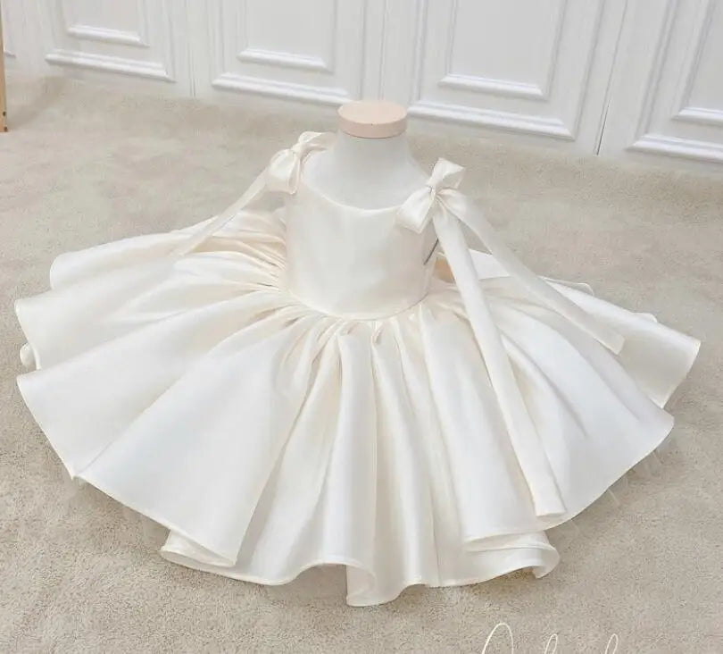 A cream-colored toddler party dress displayed on a mannequin torso. The dress features a sleeveless bodice with shoulder bows and a voluminous, tiered ruffled skirt, set against a neutral beige carpet and white paneled wall background.